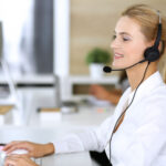 Blonde business woman using headset for communication and consulting people at customer service office. Call center. Group of operators at work at the background