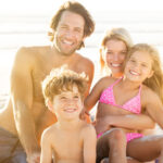 Portrait of smiling little boy with his family sitting in background at beach. Parents with children enjoying a holiday at the sea.