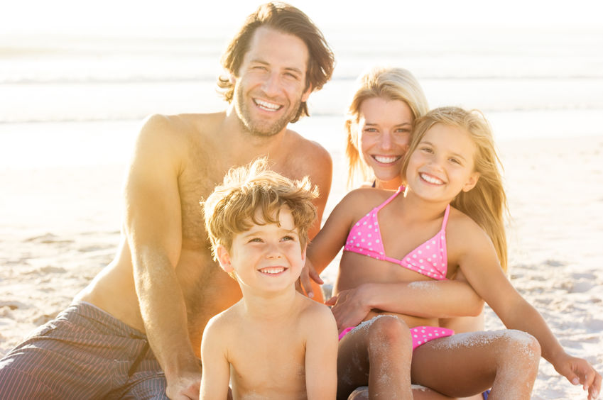 Portrait of smiling little boy with his family sitting in background at beach. Parents with children enjoying a holiday at the sea.