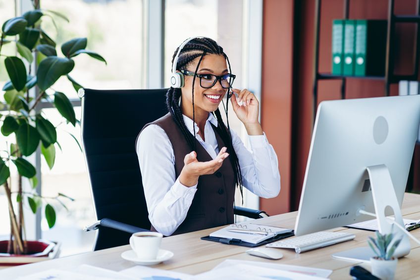young woman with glasses answering the phone with a headset on, smiling, at a computer